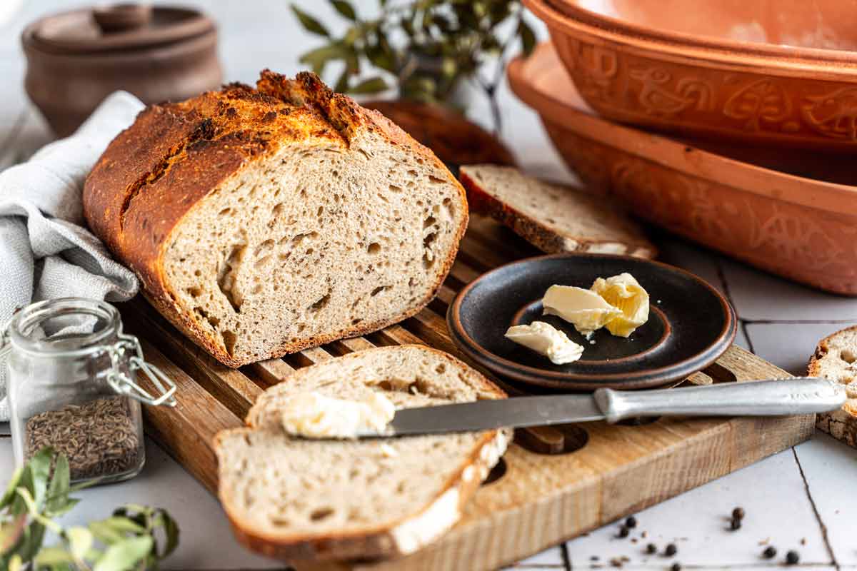 Ein angeschnittenes Brot auf einem Holzbrett, mit Buttermesser, Butter und Gewürzglas in der Nähe.