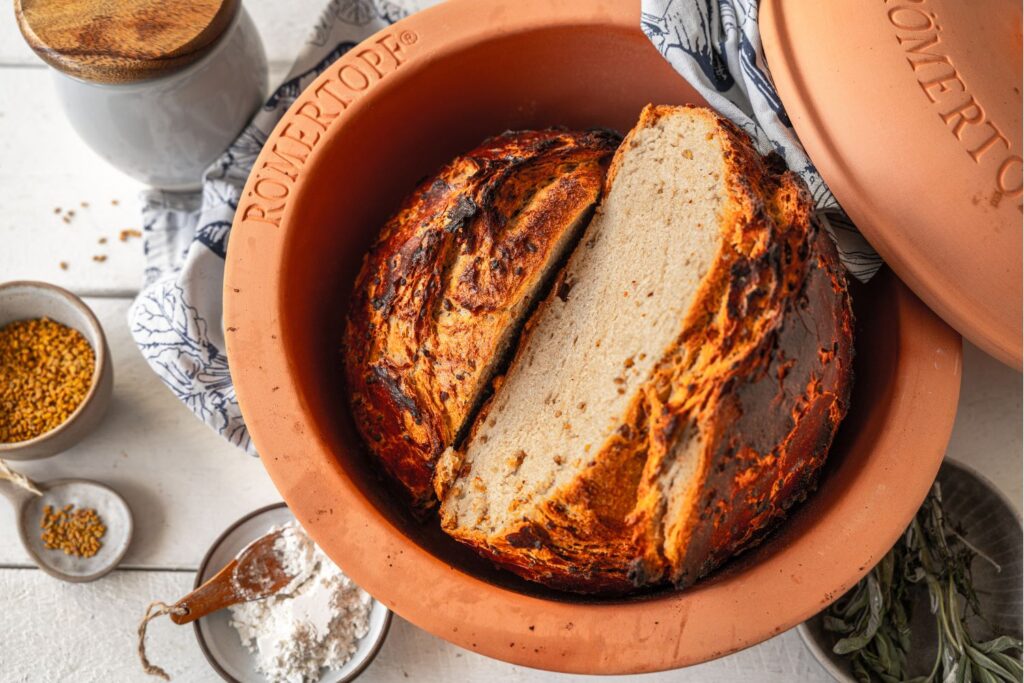 Frisch gebackenes Brot in einem runden, braunen Tontopf. Daneben liegen Gewürze und Mehl auf einem weißen Tisch.