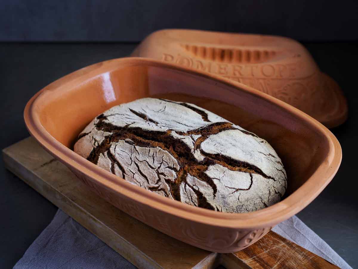 Laib Roggensauerteigbrot im geöffneten Römertopf auf einem Holzbrett
