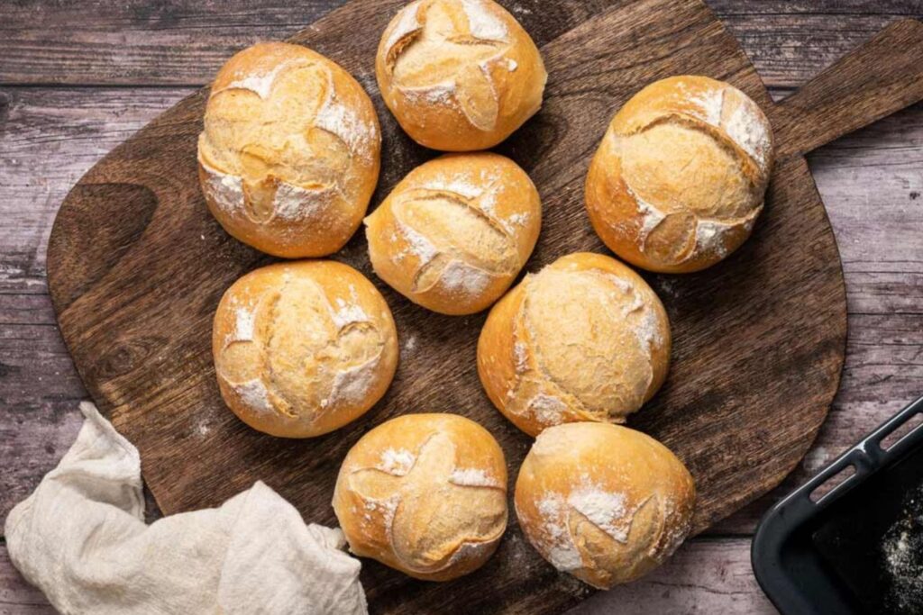Acht frisch gebackene Brötchen auf einem Holzbrett, bestäubt mit Mehl, daneben ein Geschirrtuch.