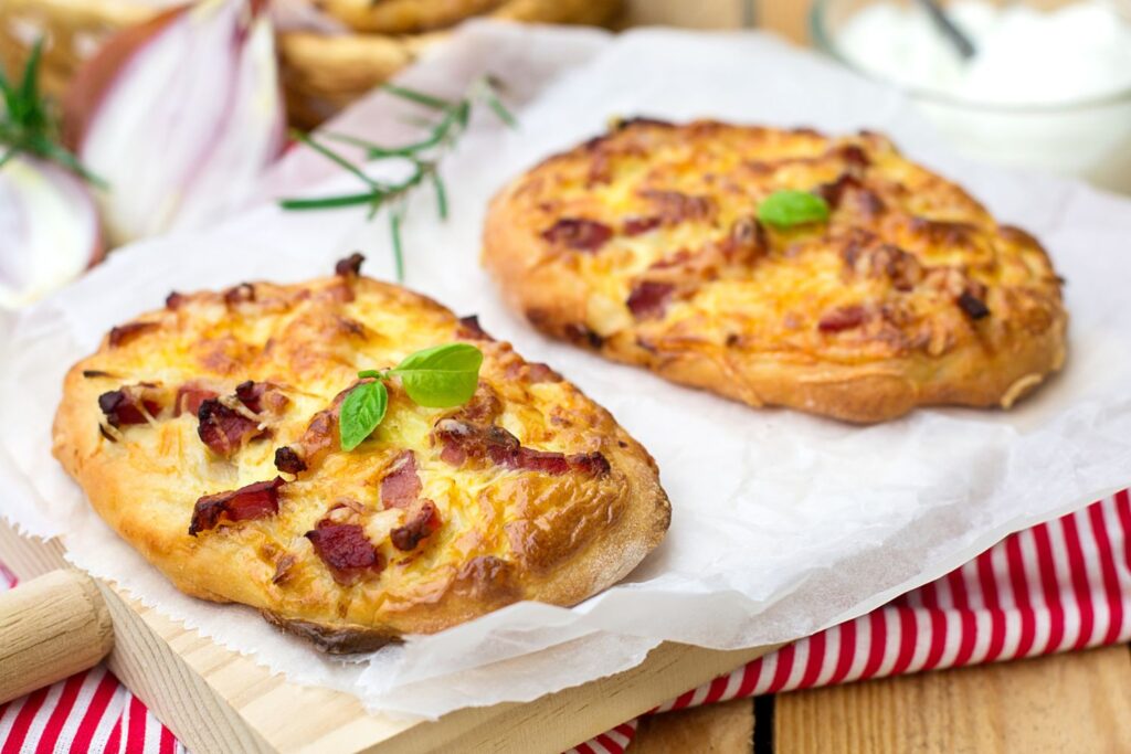 Zwei goldbraune Flammkuchen mit Speck und Zwiebeln auf Backpapier, garniert mit frischem Basilikum, auf Holzbrett.