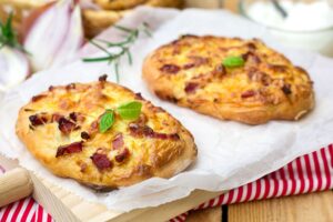 Zwei goldbraune Flammkuchen mit Speck und Zwiebeln auf Backpapier, garniert mit frischem Basilikum, auf Holzbrett.