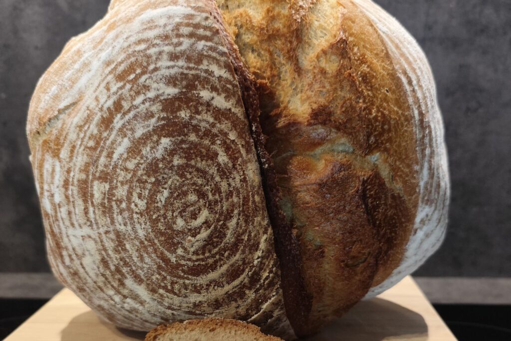 Krustiges, rundes Sauerteigbrot mit auffälligem Rillenmuster, aufgeschnitten auf einem Holzbrett vor dunklem Hintergrund.