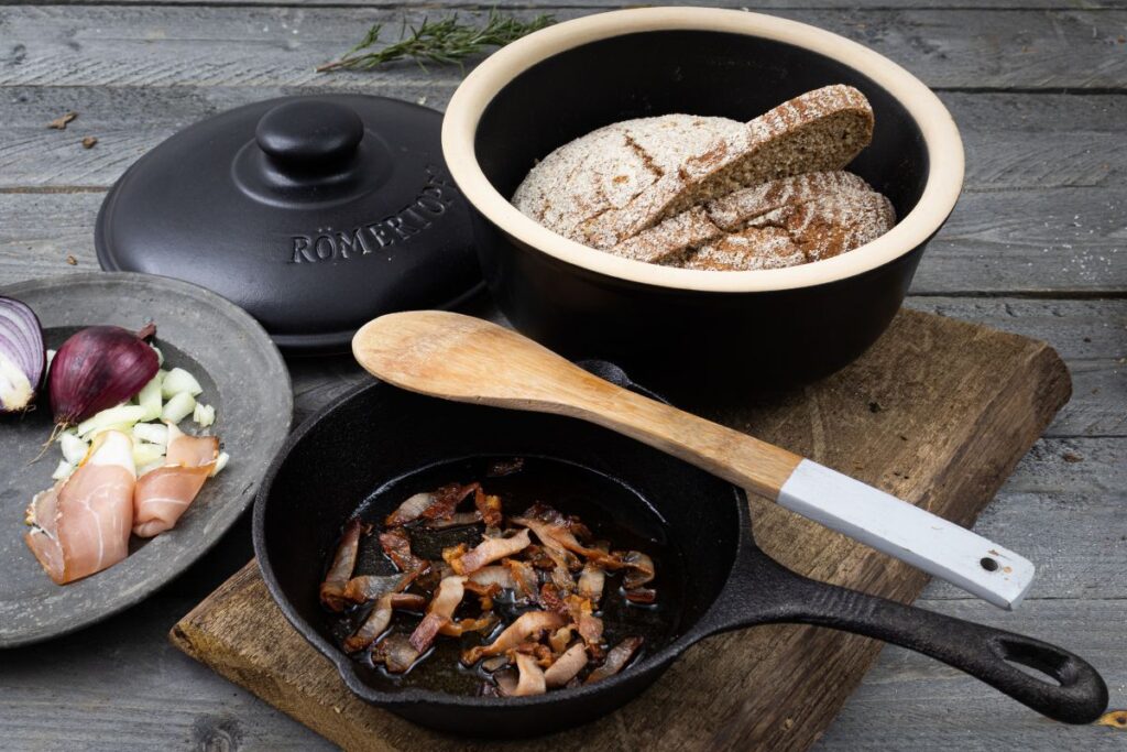 Gusseisentopf mit Brot und Speck, daneben Zwiebel und Schinken auf Holzbrett, daneben Pfanne mit gebratenem Speck.