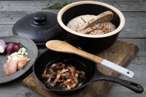 Gusseisentopf mit Brot und Speck, daneben Zwiebel und Schinken auf Holzbrett, daneben Pfanne mit gebratenem Speck.