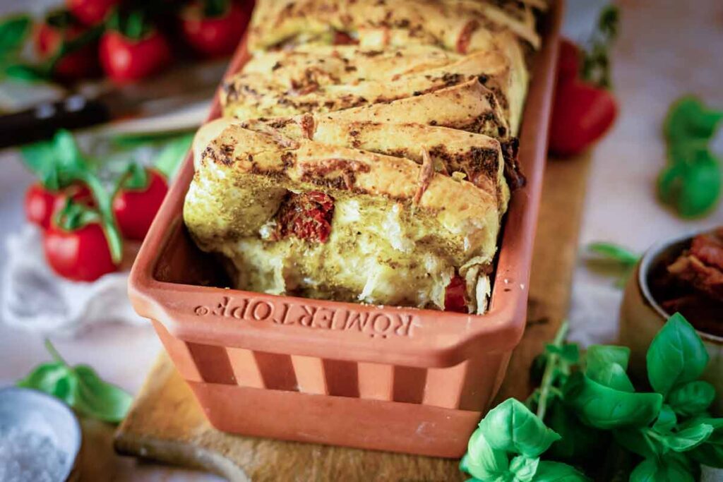 Kruste eines Brotteigs mit Gewürzen und Tomaten in einer Terrakotta-Form, im Hintergrund Basilikum und Tomaten.