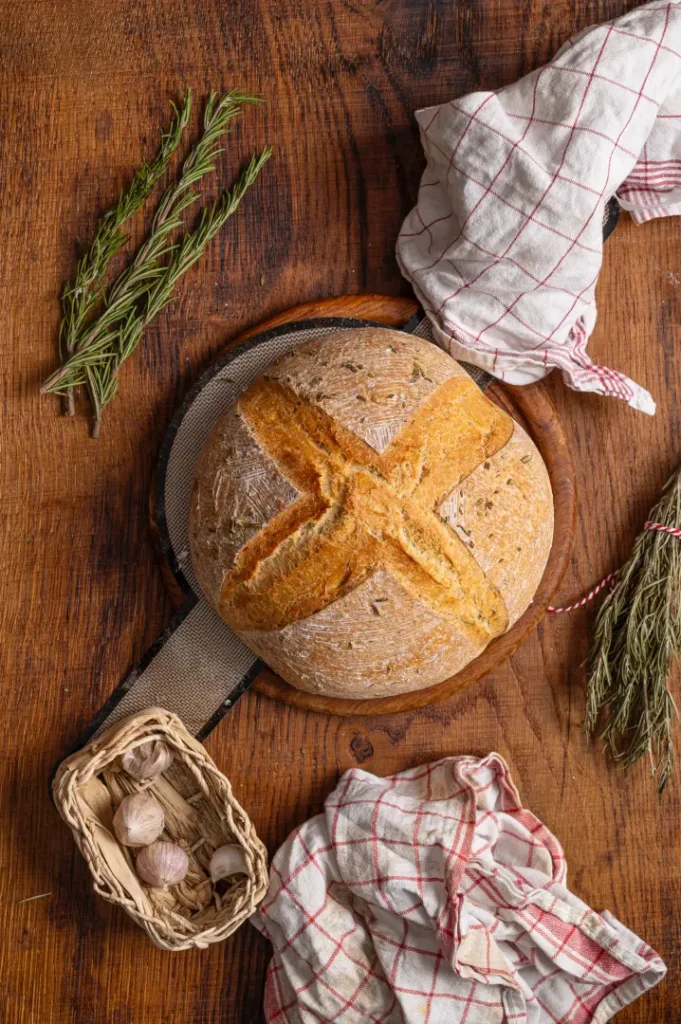 Rundes, frisch gebackenes Brot mit Kreuzmuster auf Holzbrett, umgeben von Knoblauch, Kräutern und Geschirrtüchern.