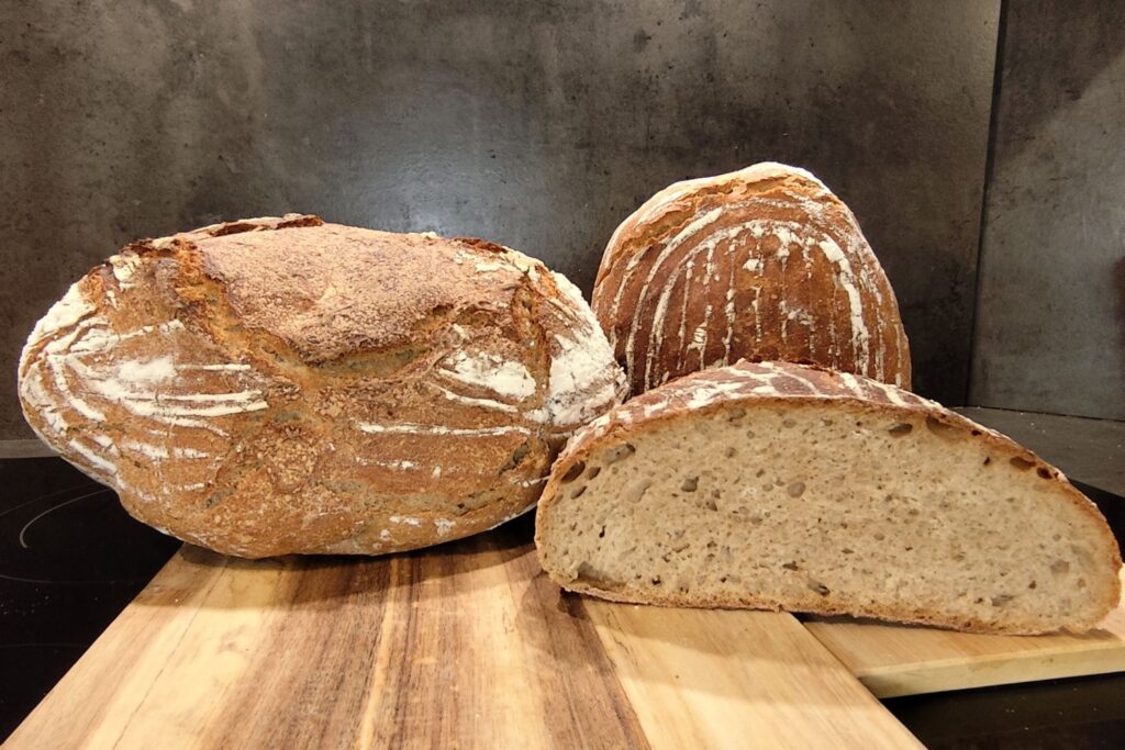 Drei Laibe frisches Sauerteigbrot auf einem Holzbrett, einer davon im Anschnitt, zeigen die grobporige Struktur.