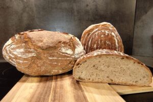 Drei Laibe frisches Sauerteigbrot auf einem Holzbrett, einer davon im Anschnitt, zeigen die grobporige Struktur.