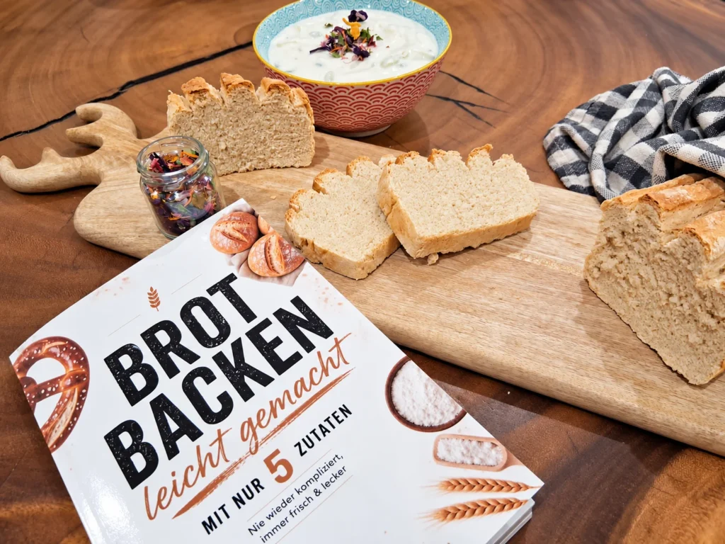 Buch „Brot backen leicht gemacht: Mit nur 5 Zutaten“ vor einem aufgeschnittenen Brot auf einem Holzbrett
